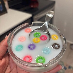 May include: A clear glass ashtray filled with a white liquid and colorful cereal pieces. A silver spoon rests in the liquid. The cereal pieces are various colors, including pink, green, yellow, and blue.