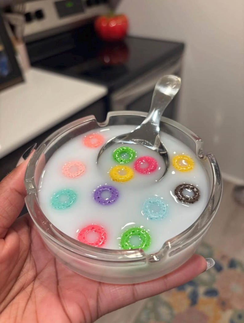 May include: A clear glass ashtray filled with a white liquid and colorful cereal pieces. A silver spoon rests in the liquid. The cereal pieces are various colors, including pink, green, yellow, and blue.