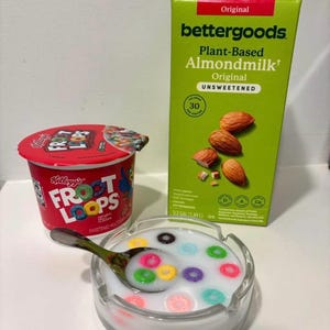 May include: A still life featuring a bowl of cereal with colorful loops in milk, a carton of Bettergoods plant-based almond milk, and a Kellogg's Froot Loops cup. The almond milk carton is green with the words "Original" and "Unsweetened".