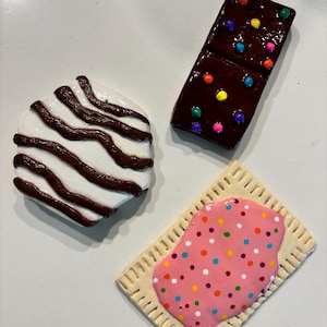 May include: Three ceramic food items: a white and brown swirled cookie, a chocolate domino with colorful dots, and a pink frosted pastry with sprinkles. The items are arranged on a white surface.