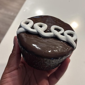 May include: A dark brown, round object with the word "eeee" in white script across the top. The object is held in a hand against a white surface. The object appears to be a decorative item.