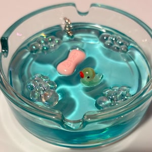 May include: A round, light blue translucent glass ashtray filled with a clear blue resin. Inside the resin are embedded decorative elements including a small pink soap bar, a tiny green rubber duck with a red beak, and clear bubble clusters.