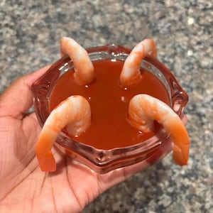 May include: A small, clear glass bowl filled with a vibrant red sauce, with four cooked shrimp arranged around the edges. The shrimp are a bright orange color, contrasting with the red sauce.