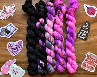 May include: Five skeins of hand-dyed yarn in shades of black, pink, and purple. The yarn is arranged on a wooden surface with several stickers featuring knitting and book-related designs. The stickers include text such as "Just One More Row."
