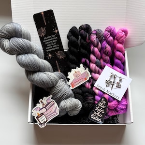 May include: A white box filled with yarn and knitting accessories. The box contains a large skein of gray yarn, several skeins of black and pink yarn, stitch markers, and stickers. One sticker reads "Fated Mates". A bookmark reads "She stitched warmth into a cold world and read herself into a brighter one."