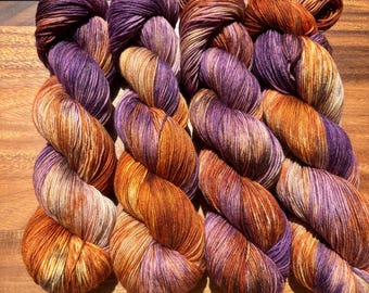 May include: Four skeins of hand-dyed yarn arranged side by side. Each skein features a variegated color palette of deep purple, burnt orange, and golden yellow tones, with visible fiber texture and twisted strands throughout the material.