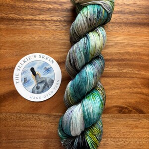 May include: A skein of hand-dyed yarn in shades of teal, green, and purple, with a cream-colored top. The yarn is twisted and sits on a wooden surface. A circular sticker with a mermaid illustration and the text "The Selkie's Skein Hand Dyed Yarn" is also present.