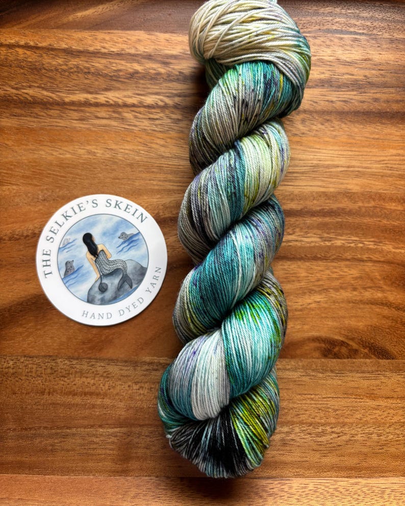 May include: A skein of hand-dyed yarn in shades of teal, green, and purple, with a cream-colored top. The yarn is twisted and sits on a wooden surface. A circular sticker with a mermaid illustration and the text "The Selkie's Skein Hand Dyed Yarn" is also present.