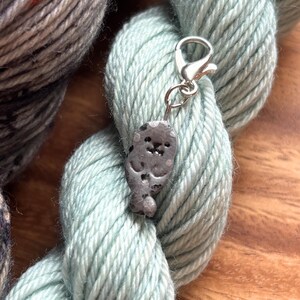 May include: A silver-colored charm in the shape of a seal is attached to a silver clasp. The charm is hanging on a light blue skein of yarn. Another skein of yarn is visible in the background. The yarn is resting on a wooden surface.