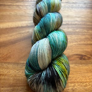 May include: A skein of hand-dyed yarn in shades of teal, green, and cream. The yarn is tightly wound, showing off the color variations. The yarn is on a wooden surface.