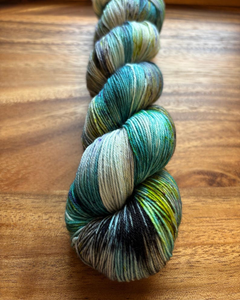 May include: A skein of hand-dyed yarn in shades of teal, green, and cream. The yarn is tightly wound, showing off the color variations. The yarn is on a wooden surface.