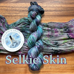 May include: A skein of hand-dyed yarn in shades of blue, green, purple, and gray. The yarn is labeled "Selkie Skin" with a circular logo featuring a mermaid. The yarn is displayed on a wooden surface.