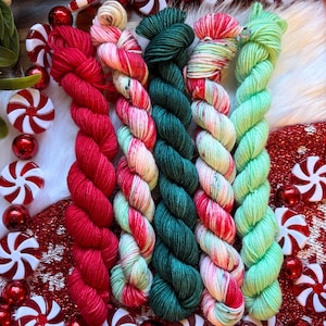 May include: Close-up of five skeins of colorful yarn in red, pink, green, and white, arranged vertically. The yarn is surrounded by red and white peppermint candies and red ornaments, creating a festive holiday theme.