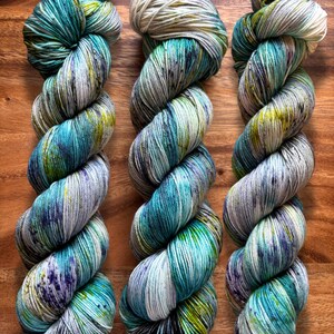 May include: Three skeins of hand-dyed yarn in shades of teal, blue, green, and purple. The yarn is twisted into a rope-like shape, with a speckled appearance. The background is a wooden surface.