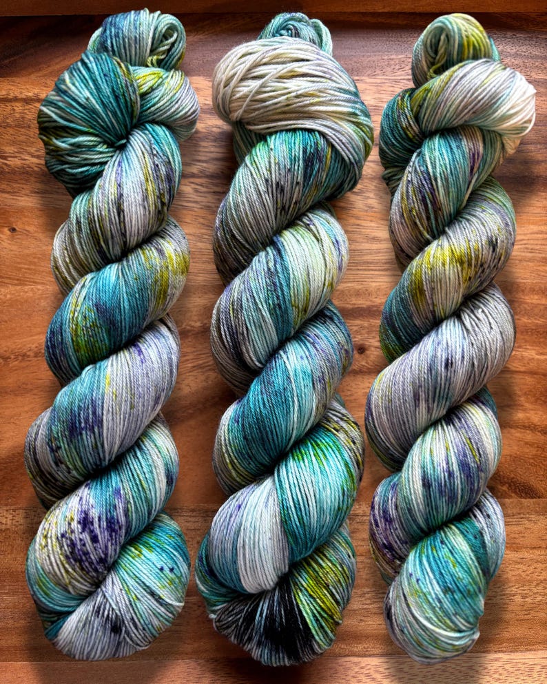 May include: Three skeins of hand-dyed yarn in shades of teal, blue, green, and purple. The yarn is twisted into a rope-like shape, with a speckled appearance. The background is a wooden surface.