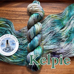 May include: A skein of hand-dyed yarn in shades of teal, green, and gray, with the word "Kelpies" in green script. The yarn is displayed on a wooden surface, with a circular label featuring a mermaid illustration.