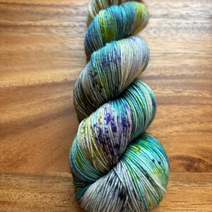 May include: A skein of hand-dyed yarn in shades of teal, blue, green, and purple. The yarn is twisted and sits on a wooden surface. The colors blend together, creating a variegated effect, perfect for knitting or crochet projects.