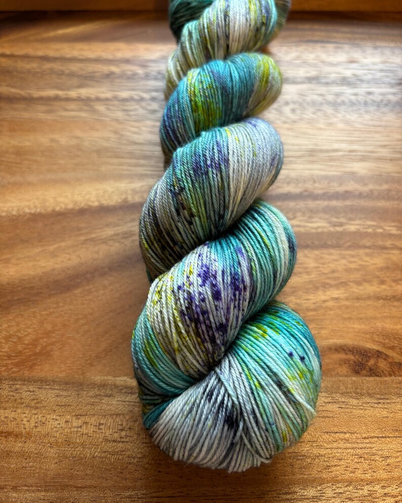 May include: A skein of hand-dyed yarn in shades of teal, blue, green, and purple. The yarn is twisted and sits on a wooden surface. The colors blend together, creating a variegated effect, perfect for knitting or crochet projects.