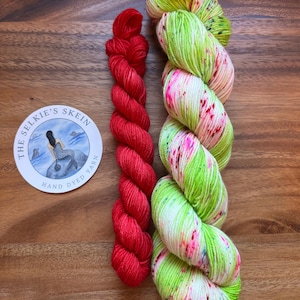May include: Two skeins of hand-dyed yarn and a round sticker on a wooden surface. One skein is a vibrant red, the other is a mix of green, white, and pink. The sticker reads "The Selkie's Skein Hand Dyed Yarn" with a mermaid illustration.