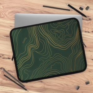 May include: A laptop sleeve with a dark green background and gold topographic lines. The sleeve has a black border and a zipper closure. The sleeve is on a wooden surface with pencils and a laptop.