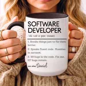 Op de afbeelding: Witte keramische mok met een zwarte handgreep en binnenkant, met de tekst "SOFTWARE DEVELOPER" en humoristische definities. De mok bevat de naam "Daniel" onderaan. Een cadeau voor softwareontwikkelaars.
