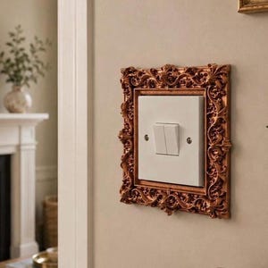 Pode incluir: Um interruptor de luz duplo branco montado numa parede, inserido numa moldura retangular decorativa de cor bronze metálico com padrões intrincados de volutas e folhas que rodeiam a placa central do interruptor.