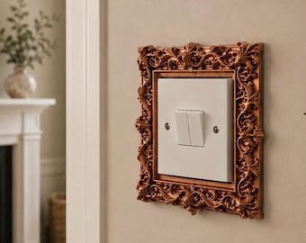May include: A white double light switch mounted on a wall, encased in a decorative, ornate, metallic bronze-colored rectangular frame featuring intricate scrollwork and leaf-like patterns surrounding the central switch plate.