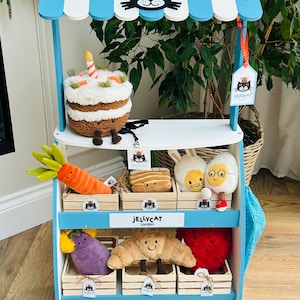 Könnte beinhalten: Ein blau-weißer Spielzeugladen-Ausstellungsstand mit dem Text "JELLYCAT GENERAL STORE" und verschiedenen Plüschtieren, darunter ein Kuchen, eine Karotte, Brot, ein Hase, ein Ei, eine Aubergine, ein Croissant und ein Herz. Der Stand hat eine gestreifte Markise.