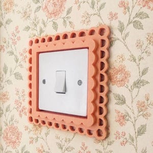 Könnte beinhalten: Eine quadratische Lichtschalterabdeckung mit einem gewellten, pfirsichfarbenen Rahmen und einem ausgeschnittenen Design. Das zentrale Element ist ein weißer Standard-Kippschalter, der mit zwei sichtbaren Schrauben an der Wand befestigt ist.