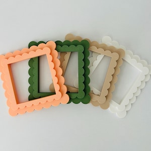 Op de afbeelding: Vier decoratieve fotolijsten in perzik, groen, beige en wit. Elk frame heeft een geschulpte rand en een vierkante opening voor een foto. De frames zijn diagonaal op een wit oppervlak geplaatst.
