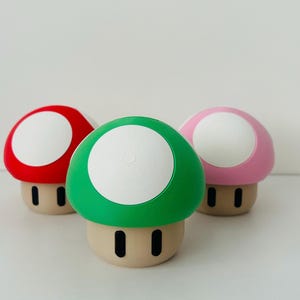 May include: Three mushroom-shaped figurines in red, green, and pink with white tops and tan bases. Each has two black rectangular eye-like details. The figurines are arranged on a white surface.