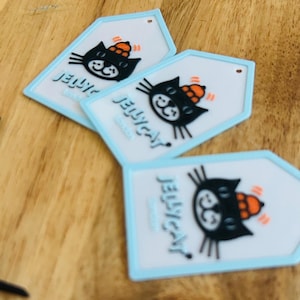 May include: A collection of white and light blue silicone tags featuring a black cat graphic wearing an orange hat. Each tag displays the text JELLYCAT London in blue lettering. The tags are shaped as squares or pentagons with a small hole at the top.
