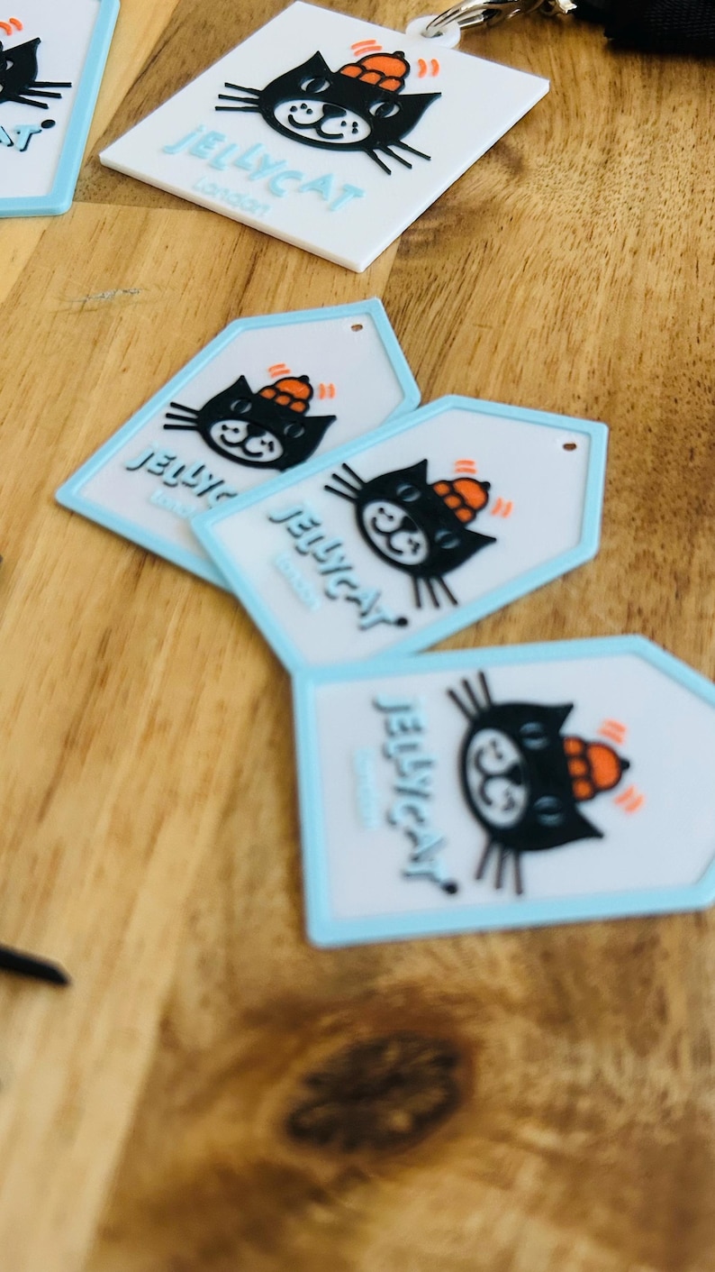 May include: A collection of white and light blue silicone tags featuring a black cat graphic wearing an orange hat. Each tag displays the text JELLYCAT London in blue lettering. The tags are shaped as squares or pentagons with a small hole at the top.