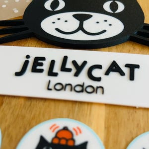 May include: A black cat-shaped emblem with a white muzzle and an orange hat, positioned above a white rectangular sign with the black text JELLYCAT London. Below, circular stickers feature the same black cat design with an orange hat.