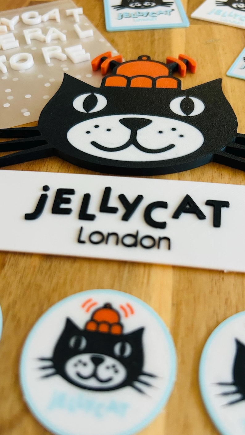 May include: A black cat-shaped emblem with a white muzzle and an orange hat, positioned above a white rectangular sign with the black text JELLYCAT London. Below, circular stickers feature the same black cat design with an orange hat.