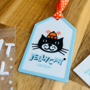 May include: A rectangular luggage tag with a light blue border and a white center. The tag features a black cat illustration wearing an orange hat, with the text JELLYCAT London printed below. An orange ribbon with white polka dots is attached.