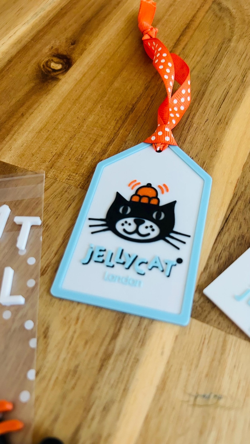 May include: A rectangular luggage tag with a light blue border and a white center. The tag features a black cat illustration wearing an orange hat, with the text JELLYCAT London printed below. An orange ribbon with white polka dots is attached.