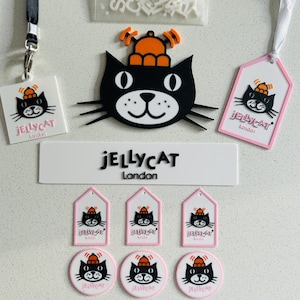 May include: A collection of Jellycat London branded items including a large black cat head with an orange hat, a rectangular sign, three house-shaped tags, three circular tags, a square tag with a black lanyard, a white tag with a ribbon, and a bag of letters.
