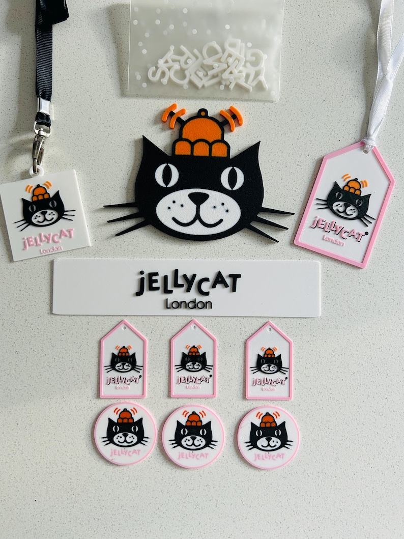 May include: A collection of Jellycat London branded items including a large black cat head with an orange hat, a rectangular sign, three house-shaped tags, three circular tags, a square tag with a black lanyard, a white tag with a ribbon, and a bag of letters.