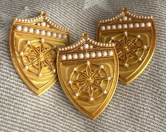 May include: Three shield-shaped pendants in a metallic gold finish. Each pendant features a central embossed compass rose design surrounded by small white spherical beads along the top edge and around the compass points, with a small loop at the top.