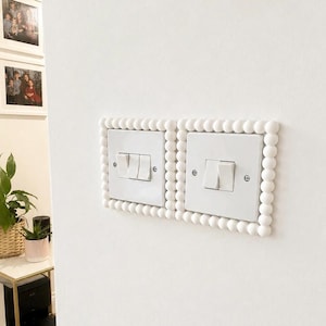 Puede incluir: Dos placas de interruptores de luz blancas montadas una al lado de la otra en una pared, cada una enmarcada por un borde decorativo de pequeñas esferas blancas uniformes. Los interruptores son de plástico blanco con tornillos plateados.