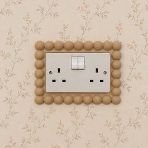 May include: A white double electrical socket with two switches, set within a decorative beige frame of small spheres. The socket is mounted on a floral wallpaper with a cream background.