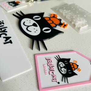 May include: A collection of Jellycat branded items including a black cat face charm with an orange hat, a white rectangular tag with the text Jellycat London, a black lanyard, and a white tag with a black cat graphic and pink border.