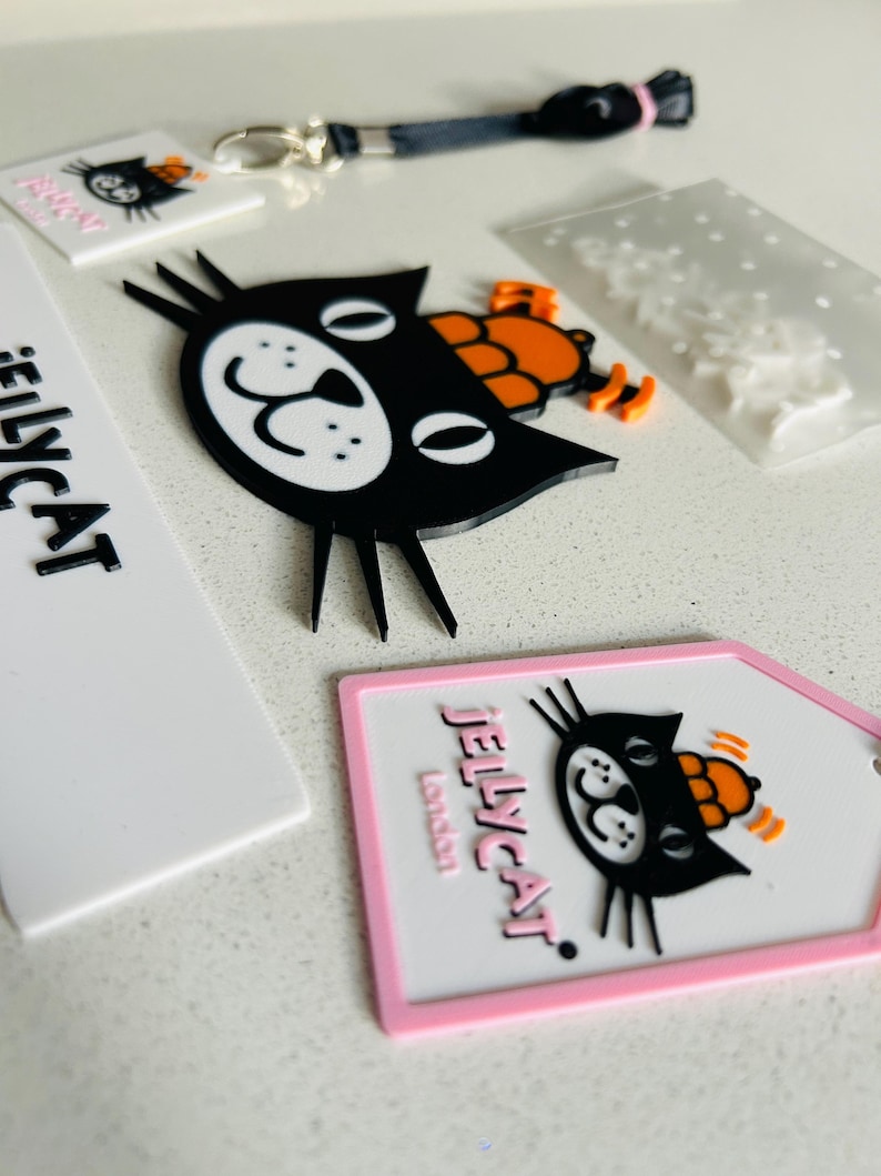 May include: A collection of Jellycat branded items including a black cat face charm with an orange hat, a white rectangular tag with the text Jellycat London, a black lanyard, and a white tag with a black cat graphic and pink border.