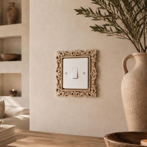 Può includere: Un interruttore della luce bianco a leva singola montato su una parete, incorniciato da una placca decorativa color beige con intricati motivi a volute e dettagli floreali intagliati che circondano la placca rettangolare e le due viti.