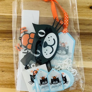 May include: A clear plastic bag containing a black cat acrylic charm with an orange ribbon, several small white rectangular stickers, and multiple light blue diamond-shaped tags. Each item features a black cat graphic with the text JELLYCAT.