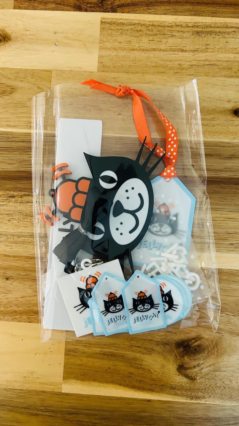 May include: A clear plastic bag containing a black cat acrylic charm with an orange ribbon, several small white rectangular stickers, and multiple light blue diamond-shaped tags. Each item features a black cat graphic with the text JELLYCAT.
