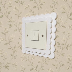 May include: A white light switch cover plate featuring a decorative scalloped edge design. The square cover surrounds a cream-coloured dual rocker light switch, which is secured by two visible silver-toned metal screws on the left and right sides.