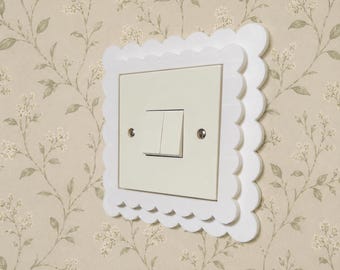 May include: A white light switch cover plate featuring a decorative scalloped edge design. The square cover surrounds a cream-colored dual rocker light switch, which is secured by two visible silver-toned metal screws on the left and right sides.