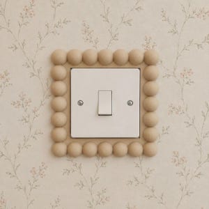 Könnte beinhalten: Ein weißer Lichtschalter mit einer hellbeigen, quadratischen Blende, die mit kleinen, erhabenen Kugeln verziert ist. Der Schalter befindet sich an einer Wand mit floralem Tapetenmuster in Beige und Rosa.